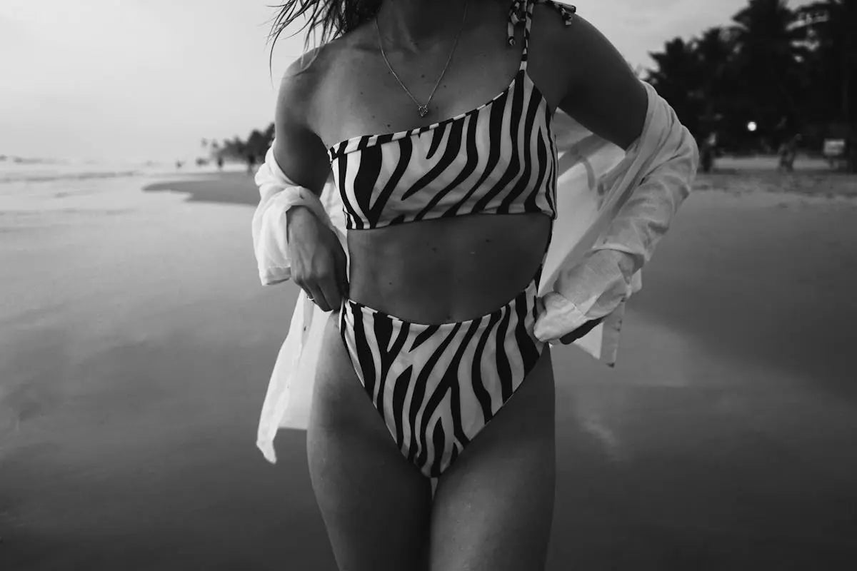 A person models a zebra-print two-piece swimsuit featuring a one-shoulder top and high-cut briefs, holding a white cover-up against a beach backdrop.