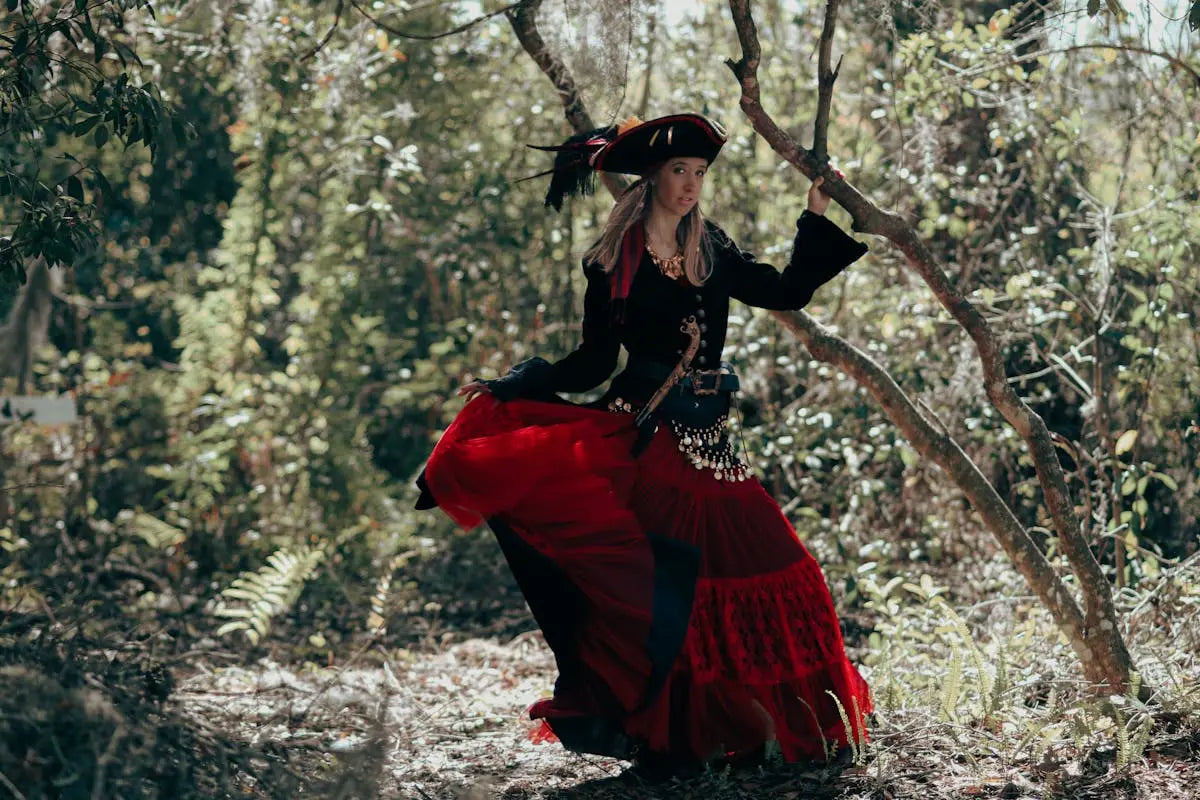 A person in a theatrical black hat with red trim and feather, black top, belt, and tiered red skirt, stands dramatically in a wooded setting.
