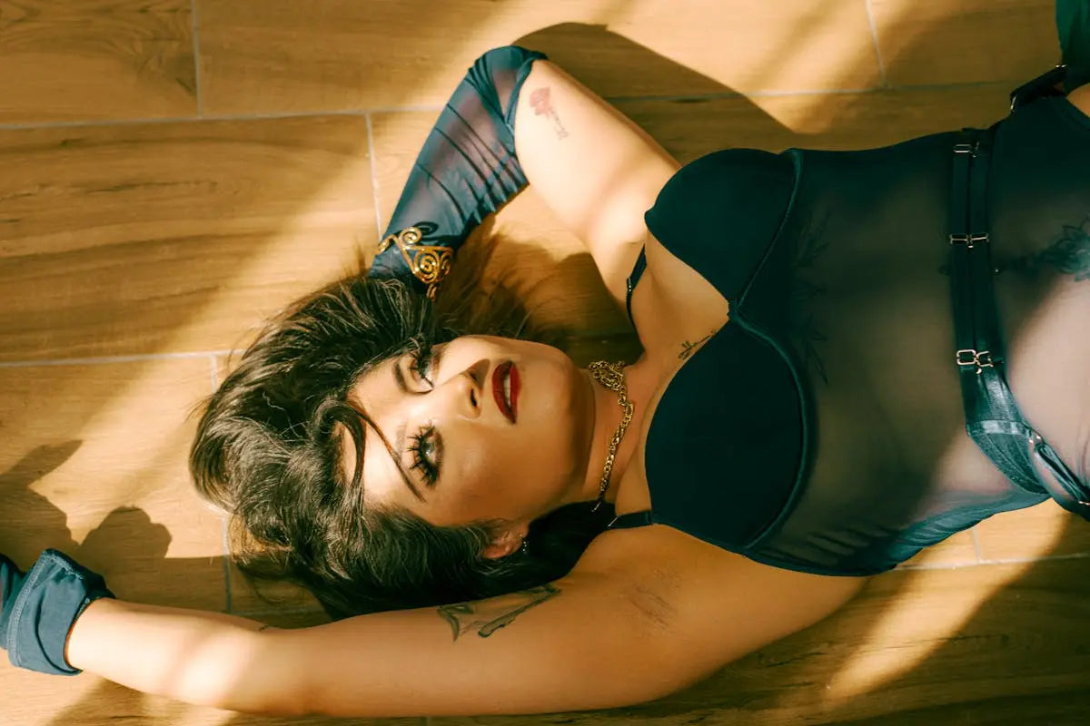 A person lies on a wooden floor wearing a black underwire bustier, sheer mesh bodysuit, mesh gloves, and gold jewelry, with tattoos visible and makeup accentuating their features.