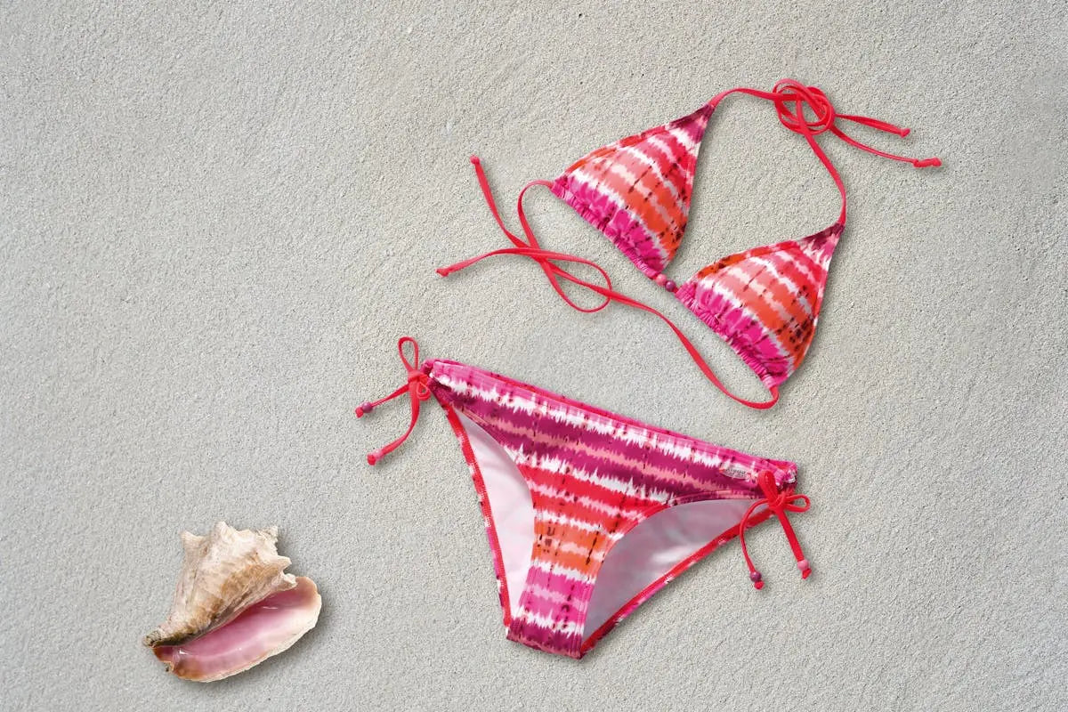 Two-piece bikini with triangle top and side-tie bottoms in pink, orange, and white tie-dye pattern, displayed on sand with a nearby seashell.