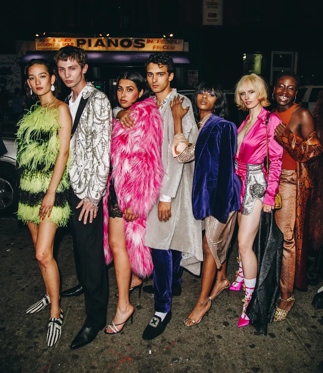 Group of seven young adults in bold eveningwear posed in front of a “PIANOS” storefront at night, showcasing vibrant fashion styles and statement pieces.