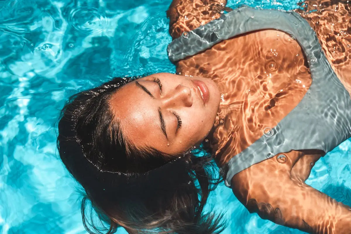 Person in a sleeveless swimsuit floats face-up in a clear blue pool with closed eyes, as sunlight creates rippling light patterns across their skin.