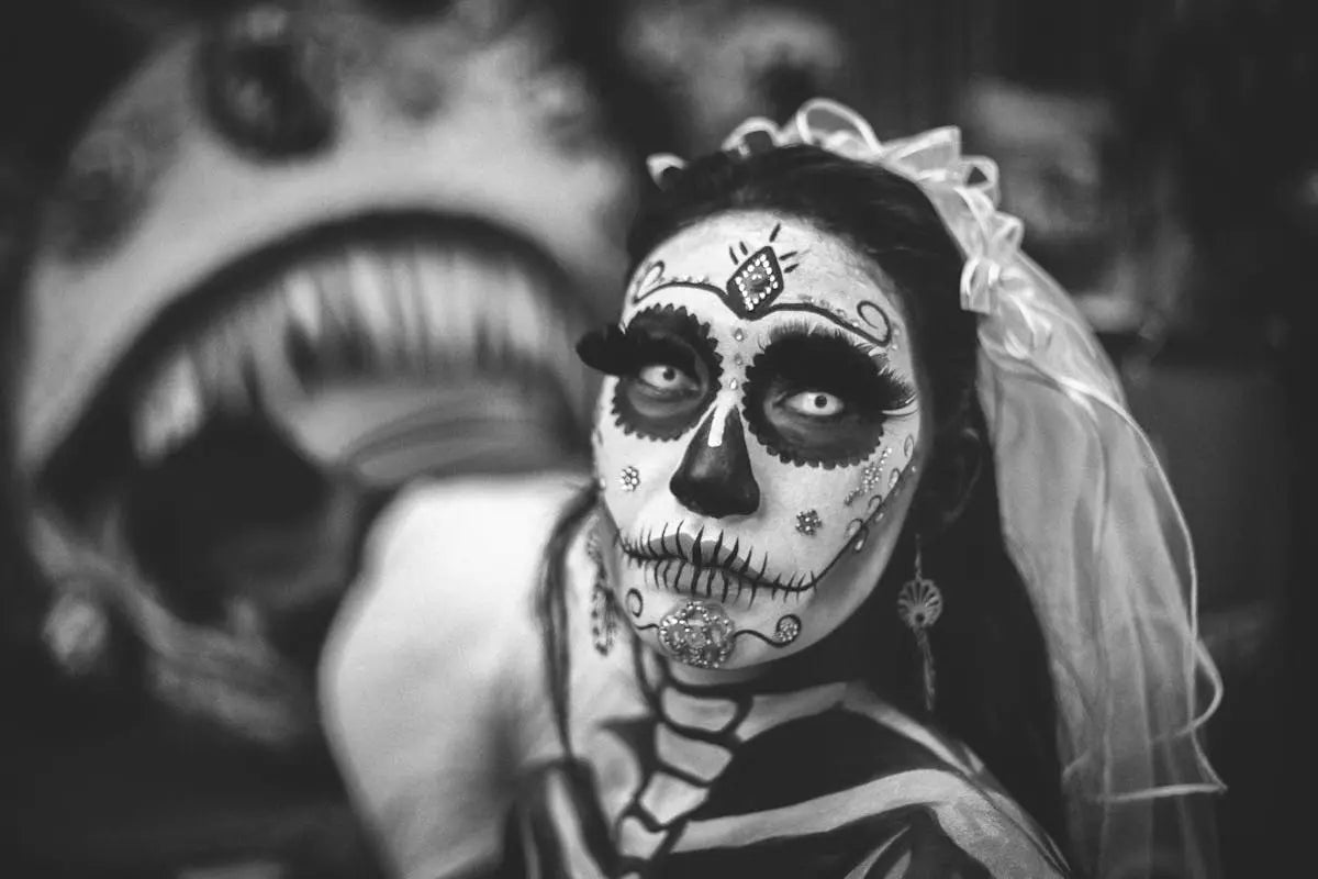 A person with Día de los Muertos sugar skull makeup featuring white face paint, dark eye circles, and skeletal rib design, wearing white contacts and a sheer veil.