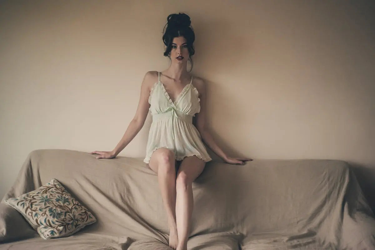 A woman in a pale camisole-style nightdress sits on a couch backrest. The couch features a plain cover and an embroidered floral-patterned throw pillow beside her.