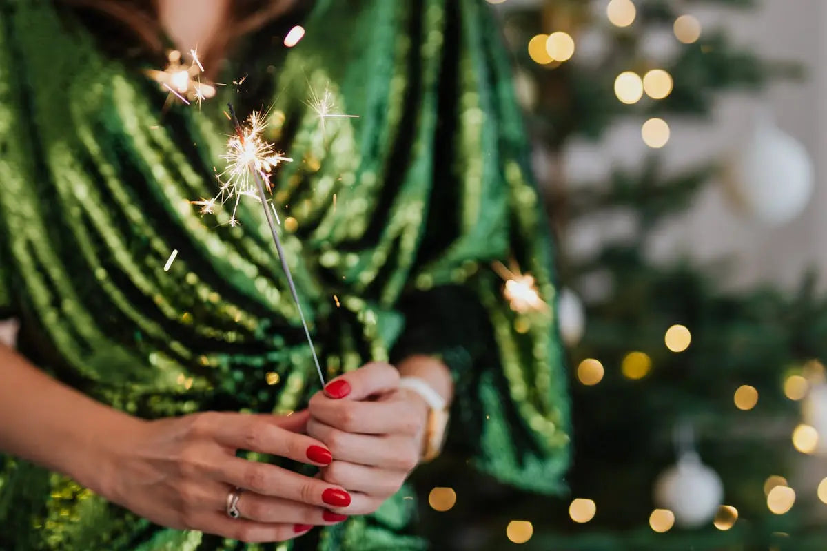 Hands holding a lit sparkler, wearing a green sequined garment with bright red nails. A softly focused Christmas tree with fairy lights and white ornaments in the background. Festive atmosphere.