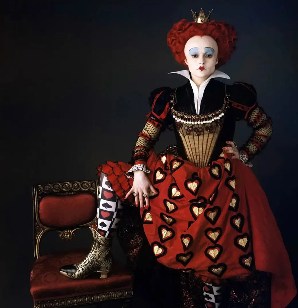 A Queen of Hearts figure in an Elizabethan gown with heart motifs, pearl-trimmed bodice, playing-card leggings, gold-embossed boots, heart-shaped red wig, and theatrical makeup.