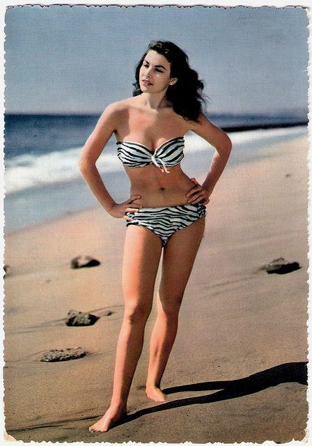 A woman wearing a bikini stands on a beach, highlighting the swimwear's design during a photo shoot.