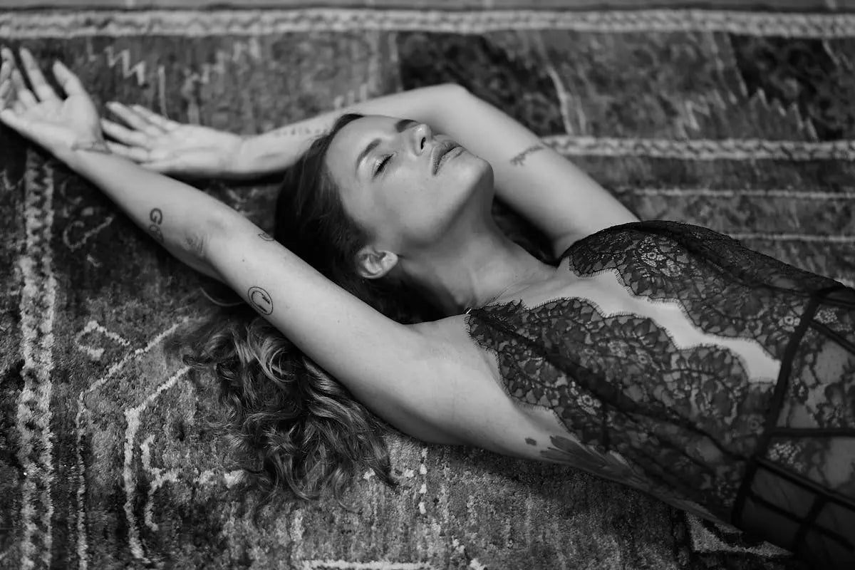 A woman wearing a black lace top is lying on a rug with her arms up, captured in a black and white photo shoot.