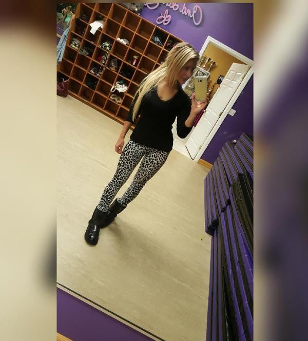 A woman in fashionable clothing and high heels takes a selfie indoors, with a curtain partially visible in the background.