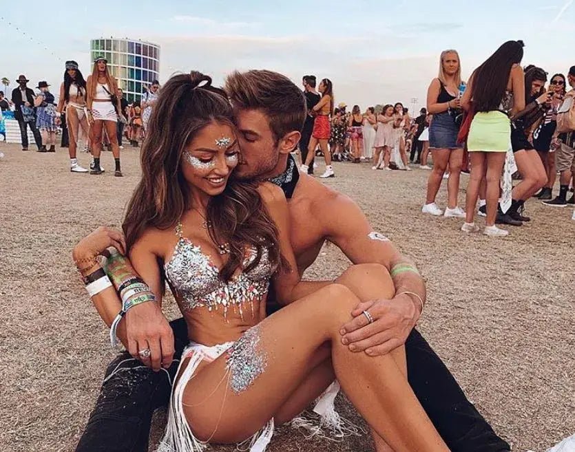 A man and woman sit on the ground among a crowd, with the woman in a short skirt, outdoors in a sunny setting.