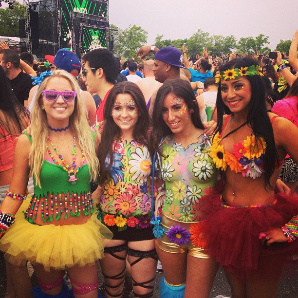 A group of women in colorful clothing at a festival, featuring body paint and flowered garments, with one man in sunglasses in the background.