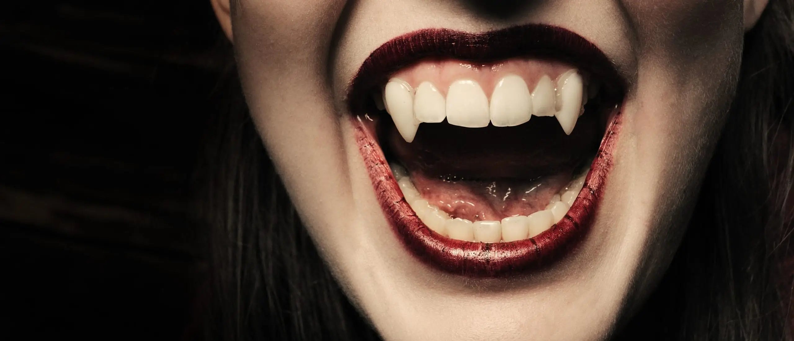 Close-up of a woman's mouth with vampire fangs, showcasing sharp teeth and glossy lips, suggesting a Halloween or costume theme.