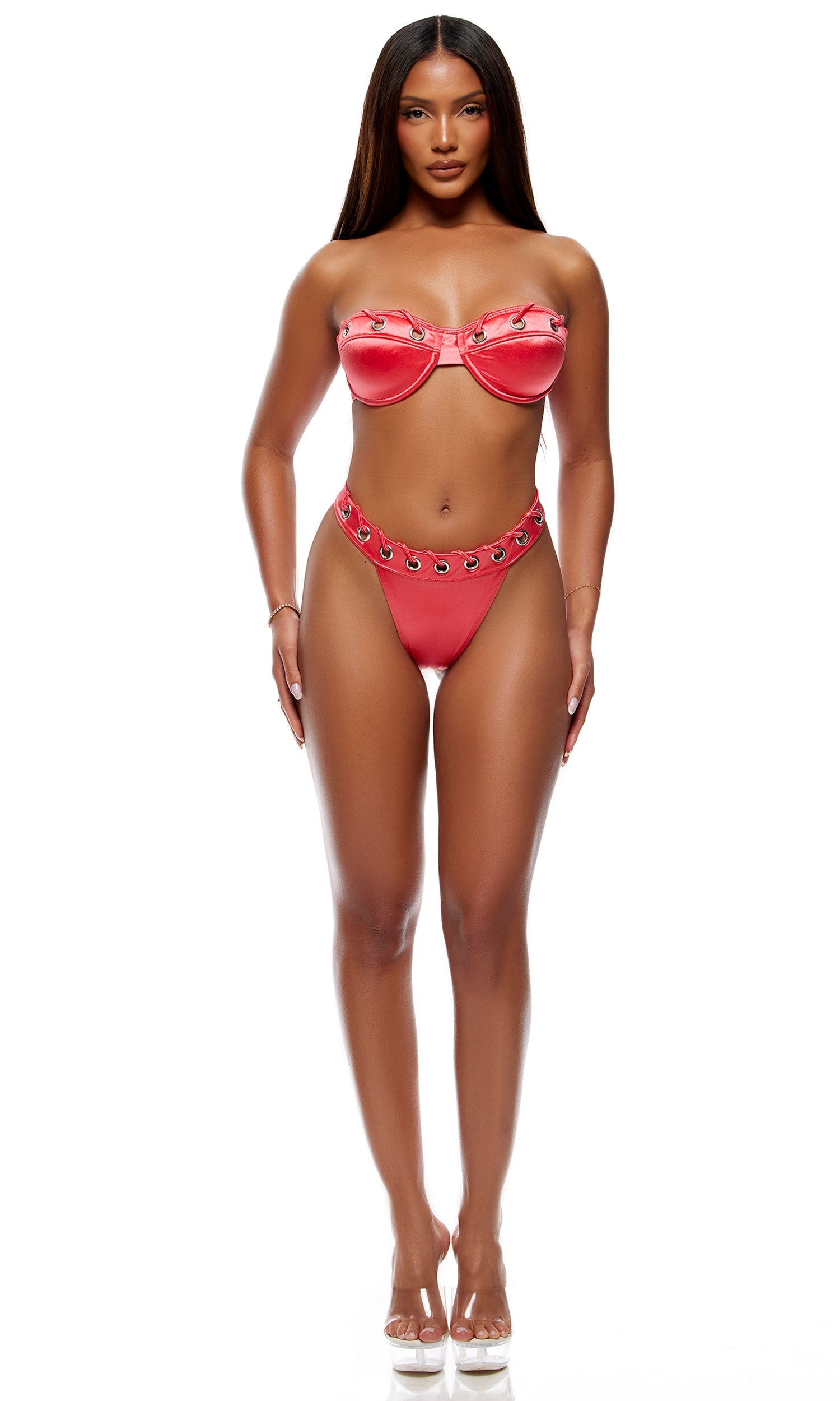 Woman modeling the Puglia Bikini Set - Coral, featuring a bandeau top with underwire whipstitch grommet cups and matching cheeky back bottoms. She wears clear-heeled sandals.