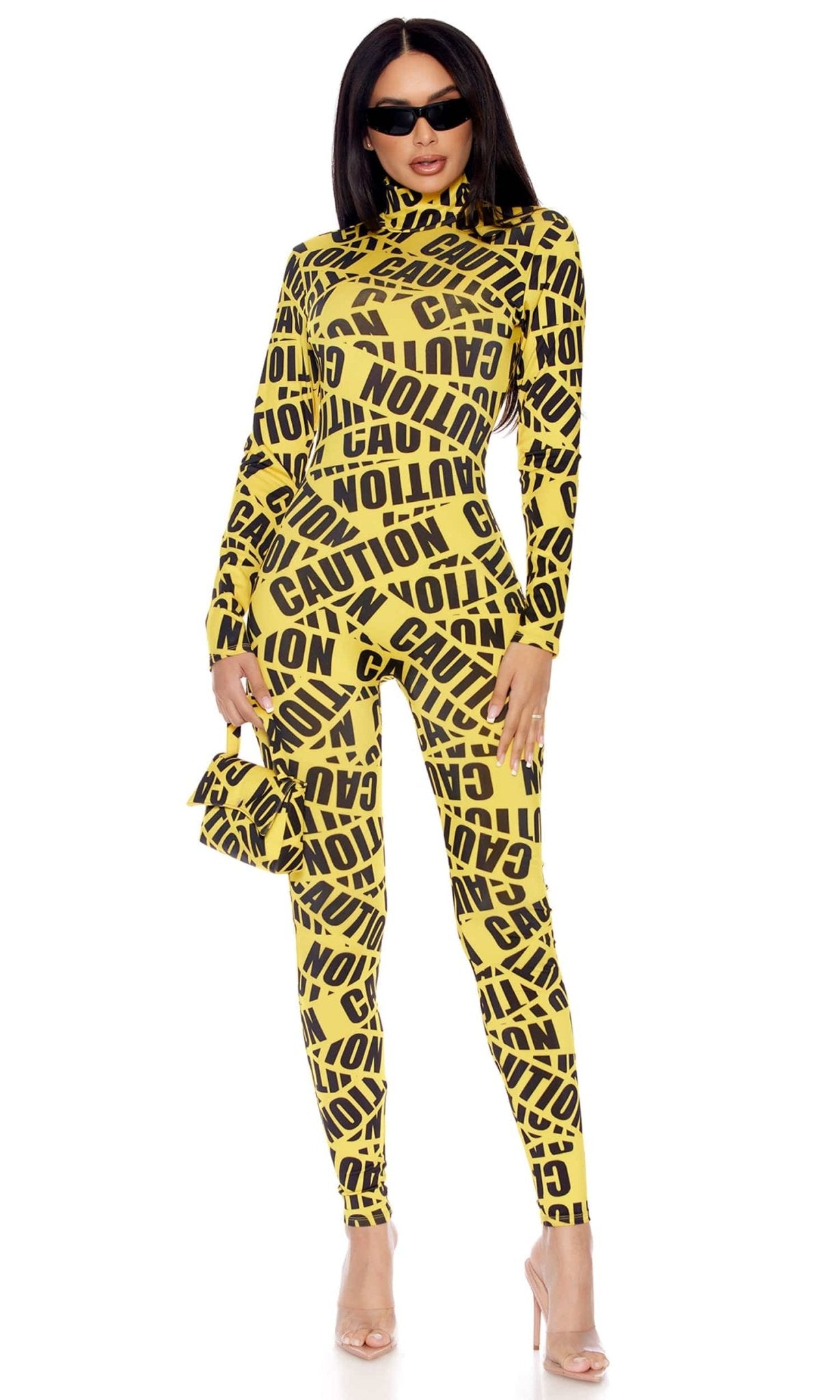 Woman in Sexy Reality TV Caution Tape Halloween Costume, featuring a mockneck jumpsuit with “CAUTION” print, sunglasses, and matching bag, posing confidently.