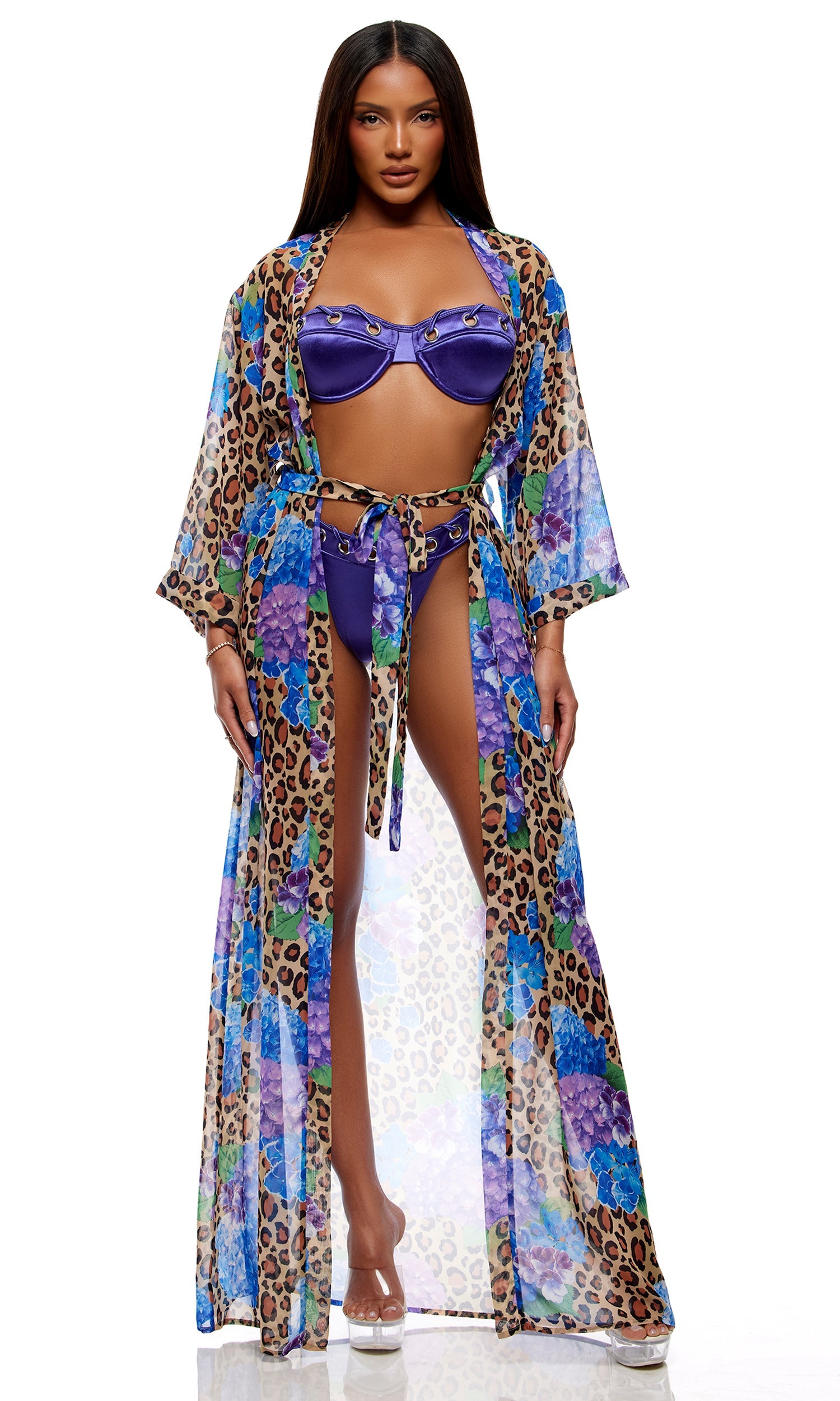 Model in Puglia Bikini Set - Purple, featuring a structured underwire top with gold-tone grommet details and matching bottoms, beneath a sheer kimono with leopard and floral print, paired with clear platform heels.